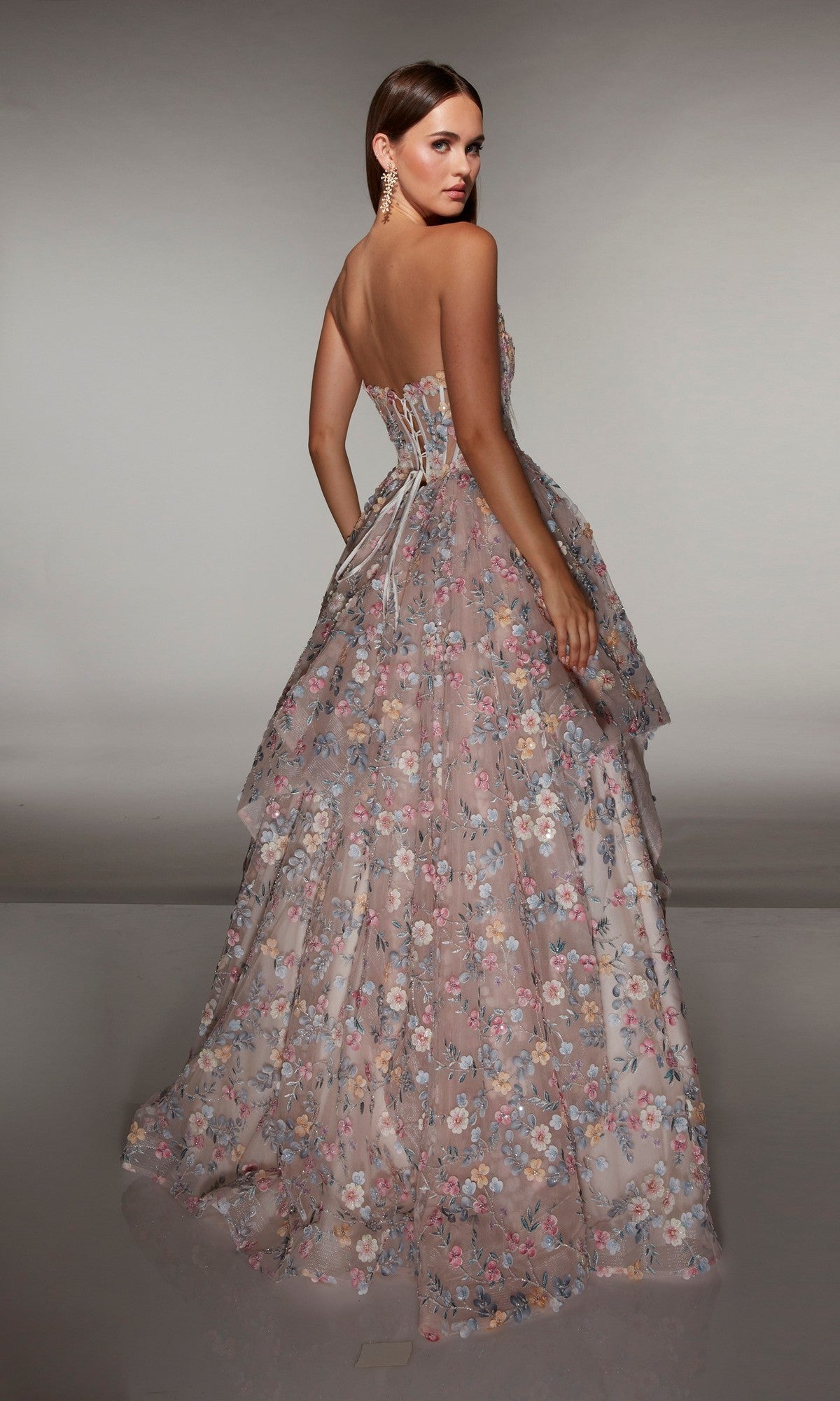 Woman wearing the Alyce 62048 long prom dress, featuring a strapless corset bodice and floral layered skirt, stands looking over her shoulder against a neutral background.